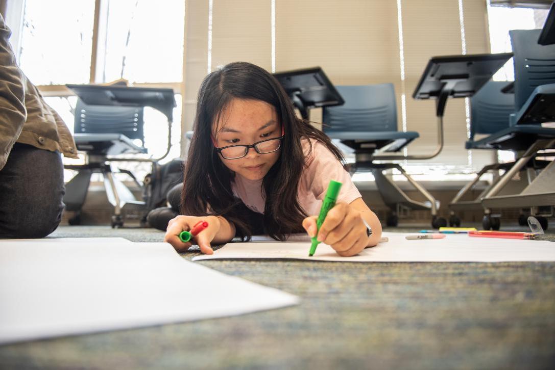 A student works on a large paper project on the floor.